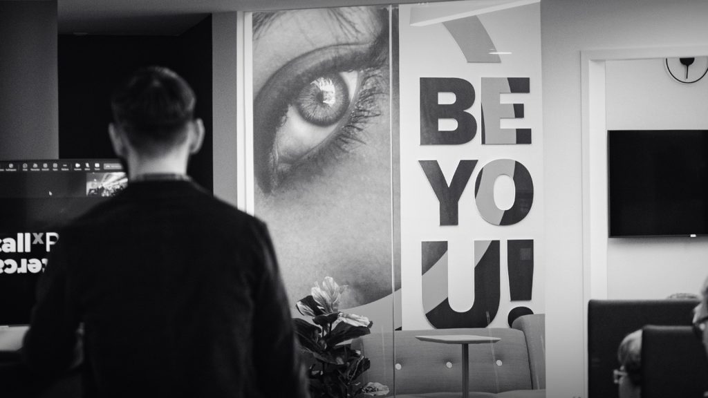 Ein Schwarz-Weiß-Foto eines BCxP-Mitarbeiters, der vor einem Schild mit der Aufschrift „BeYou“ steht.