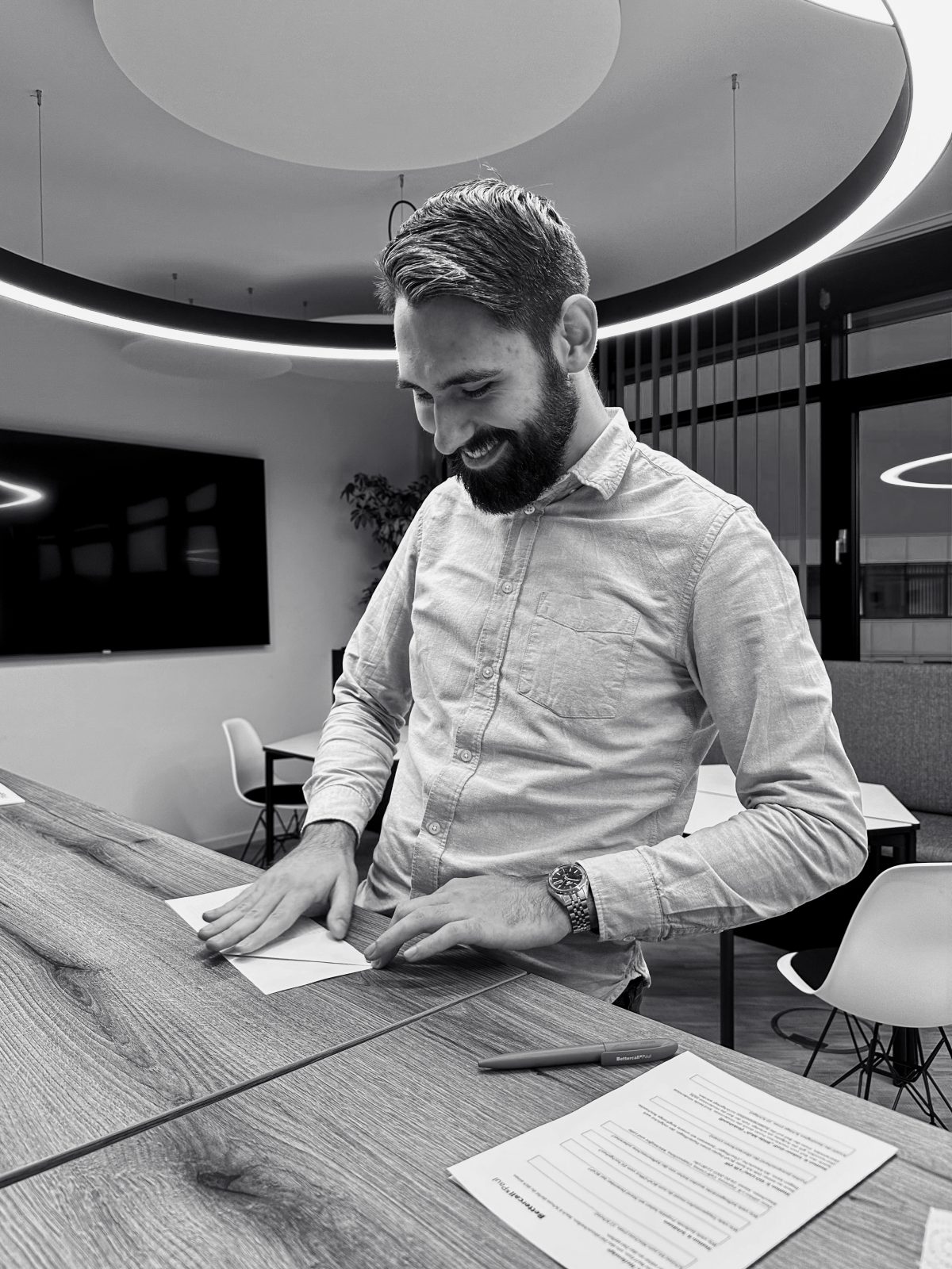 Ein Schwarz-Weiß-Foto eines lächelnden Mannes, der an einem Tisch steht und Papiere faltet.