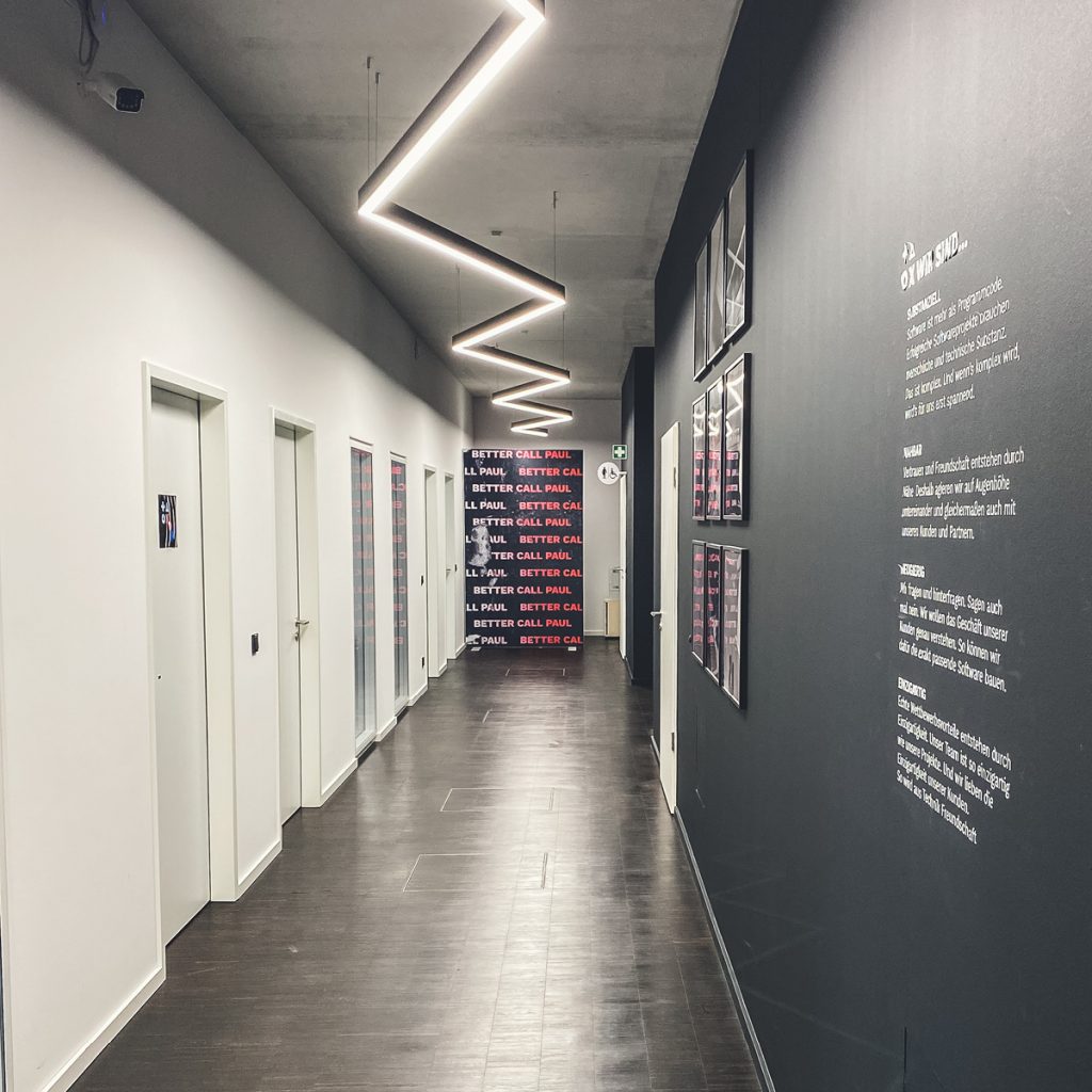 Ein moderner Korridor mit zickzackförmigen Deckenlampen, grauen Wänden mit Text und Türen auf der linken Seite.