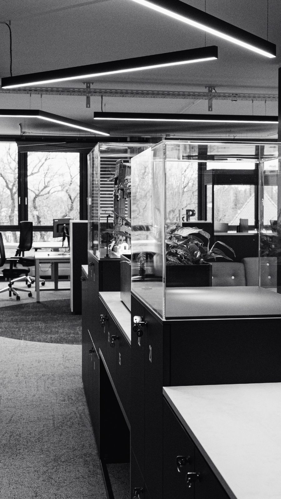 Ein moderner Büroraum mit Kabinen, Aktenschränken und Glastrennwänden, beleuchtet durch lineare Deckenleuchten.