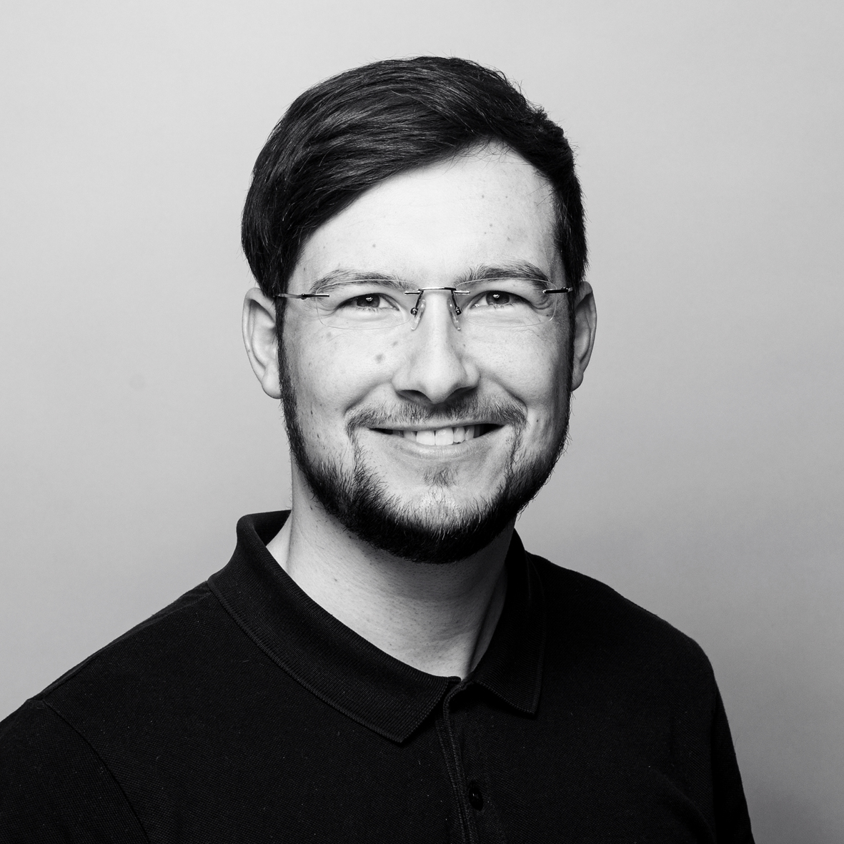 Ein Schwarz-Weiß-Foto eines jungen Mannes mit kurzem, dunklem Haar und Brille. Er trägt ein dunkel gefärbtes Polohemd und lächelt freundlich in die Kamera.