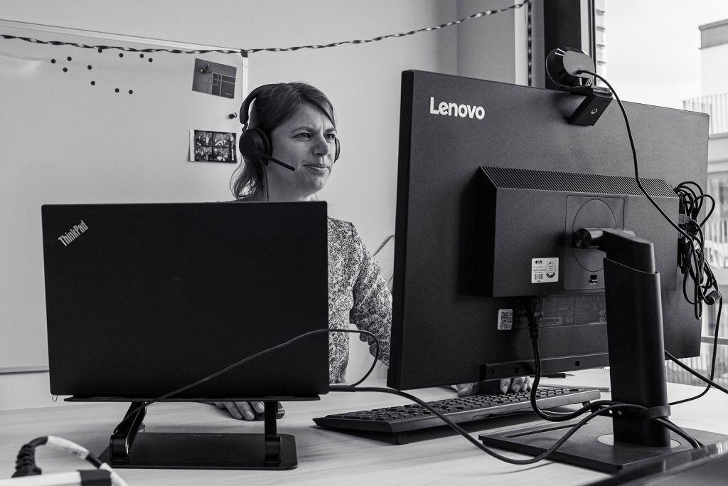 Eine Frau mit Headset sitzt an einem Schreibtisch und arbeitet konzentriert an zwei Lenovo-Bildschirmen, einer davon ein Laptop. Sie befindet sich in einem modernen Büro mit einem Fenster im Hintergrund.