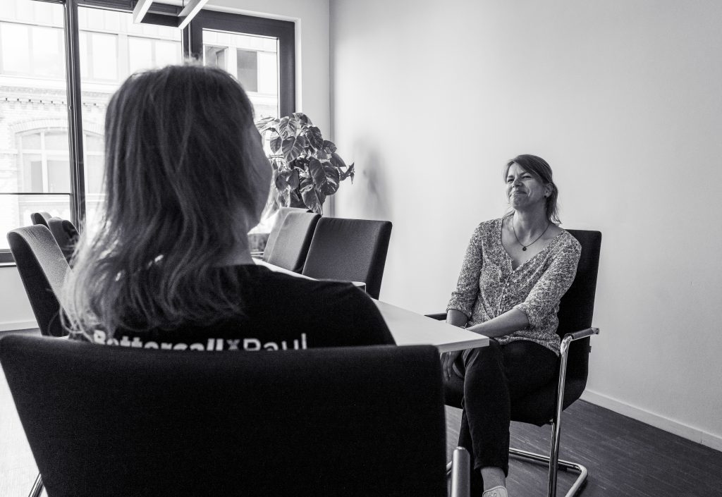 Zwei Frauen sitzen in einem Büro an einem Tisch und unterhalten sich. Eine der Frauen lächelt leicht, während sie der anderen zuhört. Im Hintergrund ist ein großes Fenster mit Blick auf ein Gebäude zu sehen.