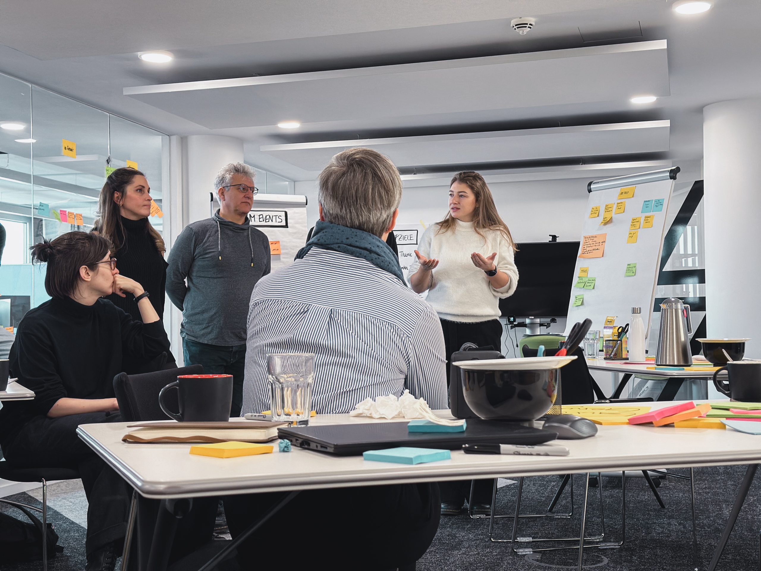 Eine Gruppe von Personen diskutiert in einem modernen Büro, mit Haftnotizen auf einem Whiteboard und Arbeitsmaterialien auf Tischen im Vordergrund.