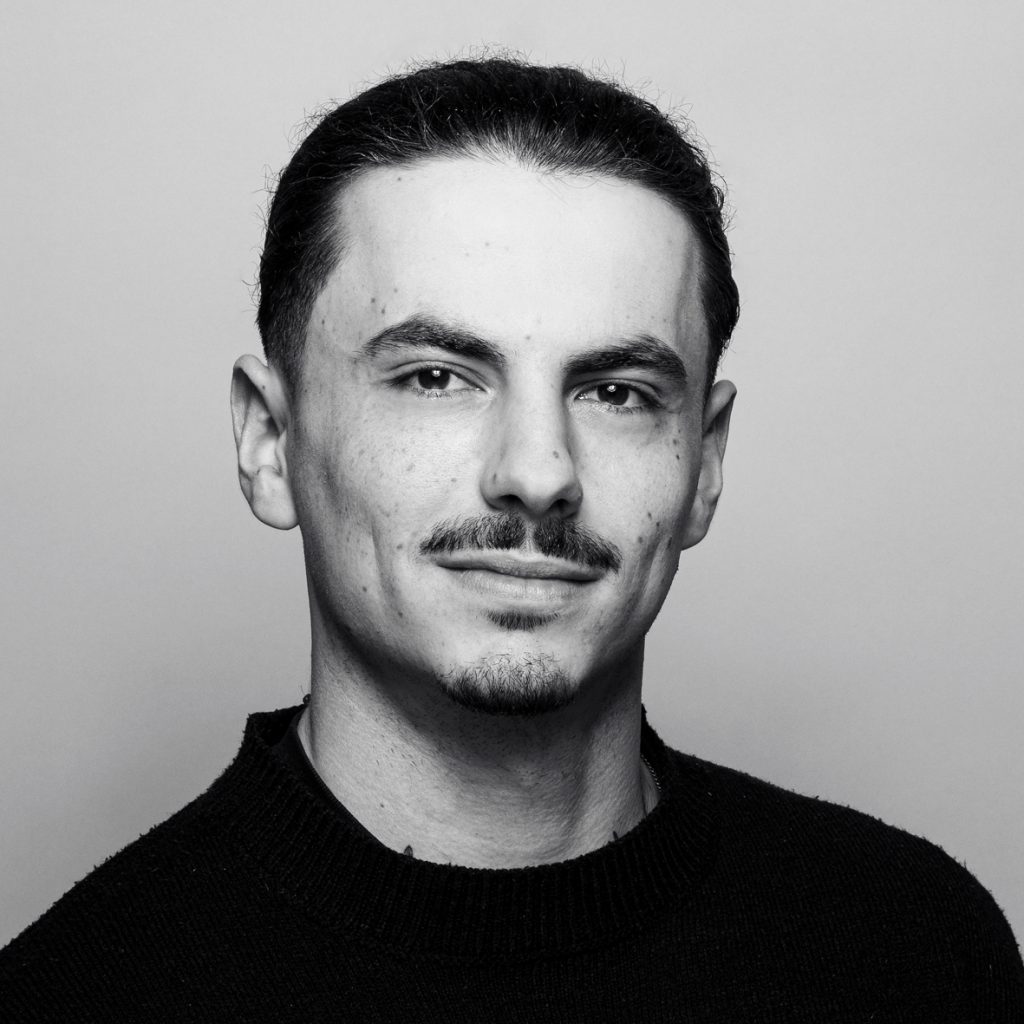 Porträtfoto eines jungen Mannes mit dunklen, zurückgekämmt gebundenen Haaren, schmalem Schnurrbart und kleinem Kinnbart. Er trägt einen dunklen Strickpullover und schaut ruhig lächelnd in die Kamera. Schwarz-weiß-Aufnahme vor hellem Hintergrund.