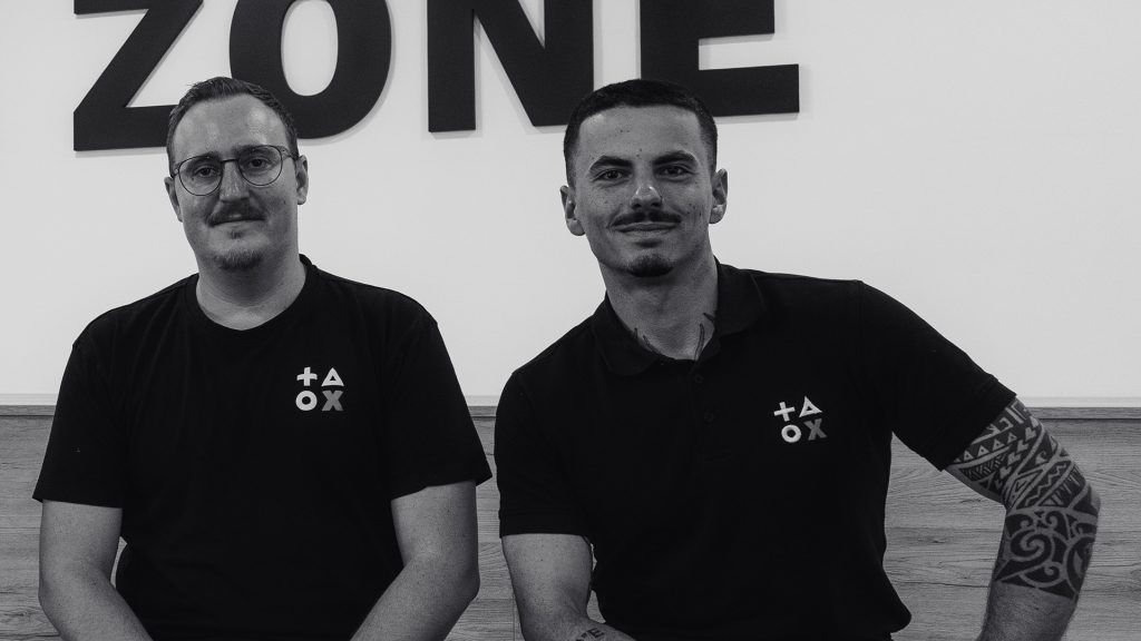 Schwarz-weiß-Aufnahme von zwei Personen, die nebeneinandersitzen. Beide tragen dunkle Shirts mit den BCxP-Buddies. Die Person links hat kurze Haare, Brille und einen leichten Schnurrbart. Die Person rechts hat kurze Haare, einen gepflegten Bart und sichtbare Tattoos am rechten Arm. Hinter ihnen sind große Buchstaben des Wortes ‚ZONE‘ an der Wand zu sehen.