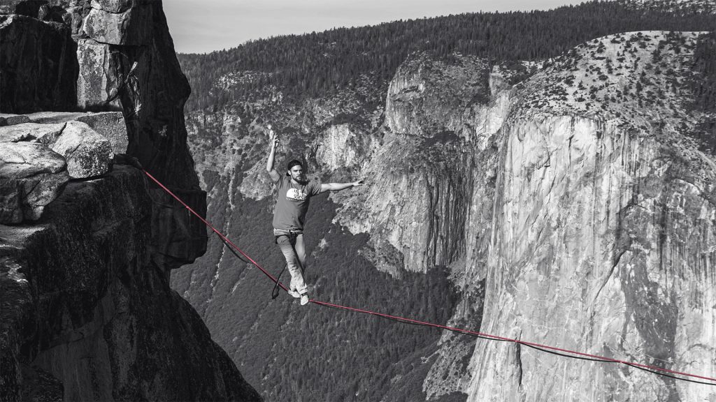 Eine Person balanciert in großer Höhe auf einem schmalen Seil zwischen zwei Felsen. Das Bild ist schwarz-weiß, nur das rote Seil ist farbig hervorgehoben. Im Hintergrund sind steile Felswände und Wälder zu sehen.
