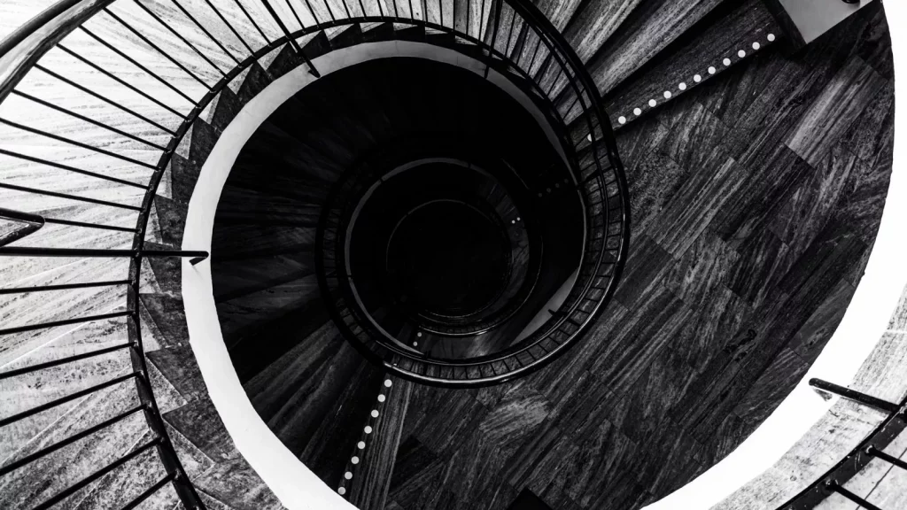 Schwarz-weiß Fotografie einer spiralförmigen Treppe mit Geländer, die sich in die Tiefe windet.