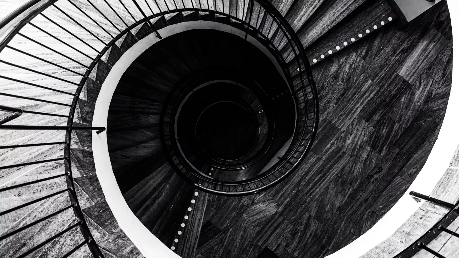 Schwarz-weiß Fotografie einer spiralförmigen Treppe mit Geländer, die sich in die Tiefe windet.