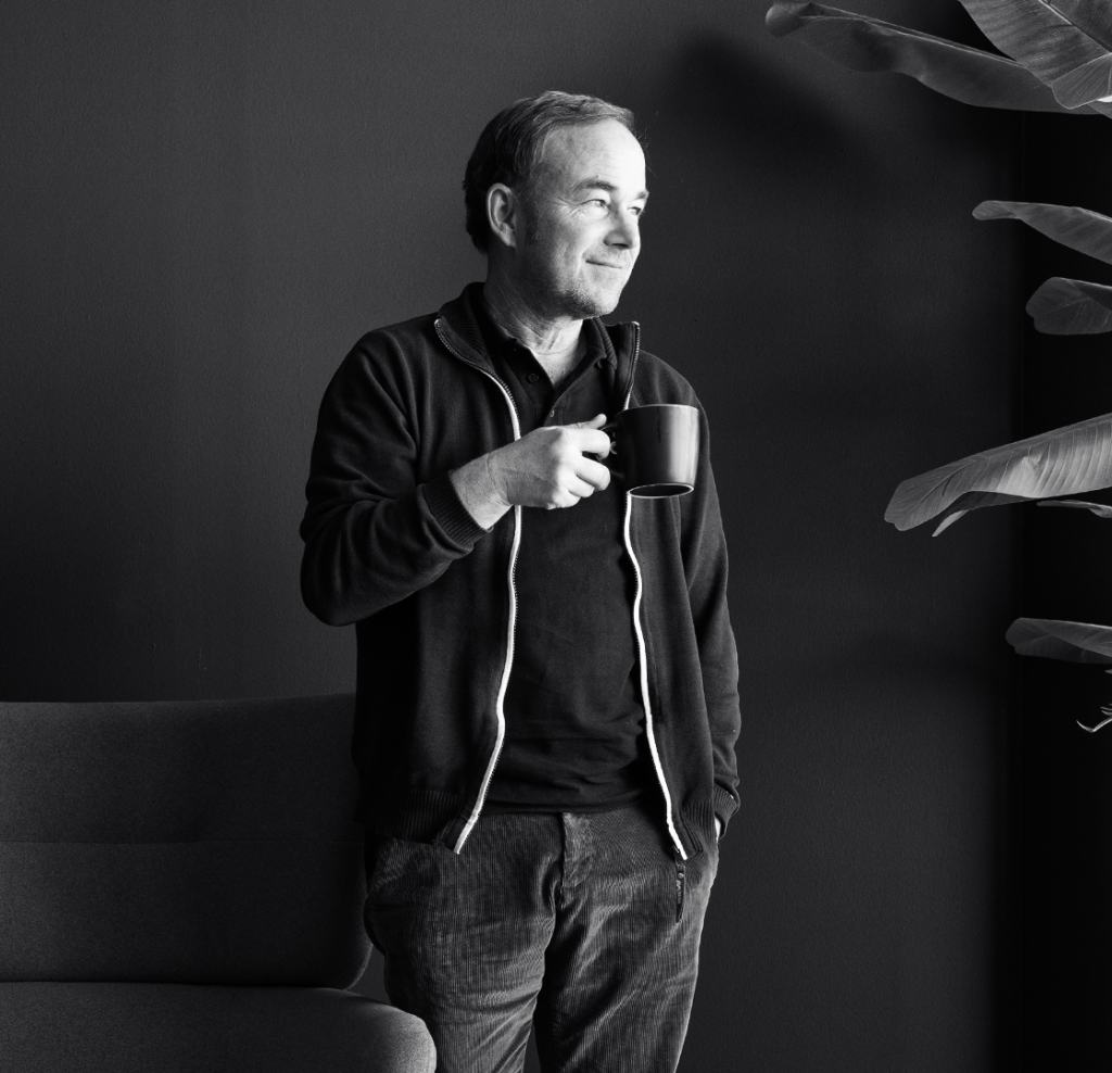 Ein Mann mittleren Alters steht entspannt mit einer Tasse Kaffee in der Hand vor einer dunklen Wand. Er blickt lächelnd zur Seite. Neben ihm stehen ein Sofa und große Pflanzenblätter, die dem schwarz-weiß aufgenommenen Bild eine ruhige, gemütliche Atmosphäre verleihen.