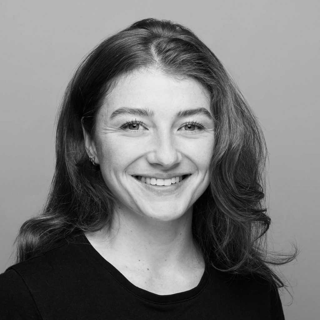 Porträt einer lächelnden Person mit schulterlangem, leicht gewelltem Haar, die direkt in die Kamera blickt. Schwarz-weißes Foto vor neutralem Hintergrund.