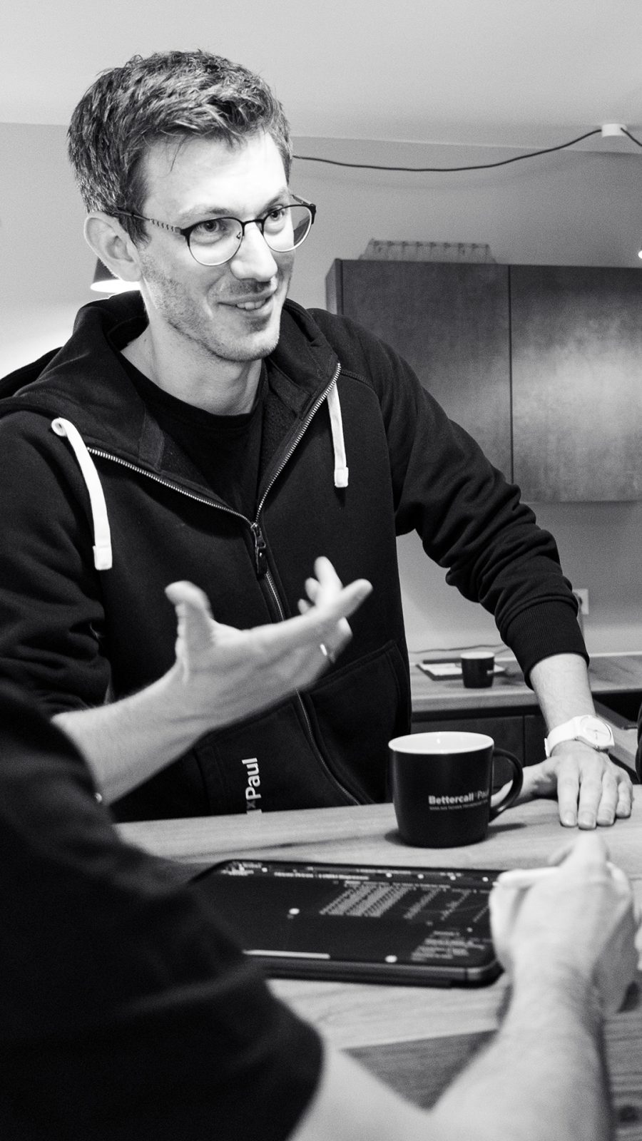 Ein Mitarbeiter mit Brille und Hoodie gestikuliert im Gespräch an einem Tisch, vor ihm steht eine Kaffeetasse, im Vordergrund liegt ein Tablet mit Code.