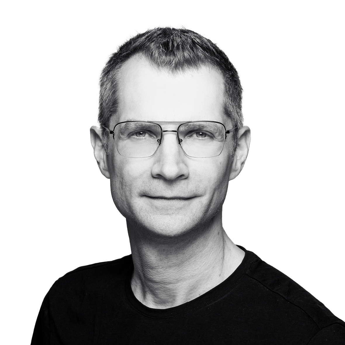 Ein Mann mit kurzen Haaren und Brille, der ein schwarzes Hemd trägt und mit neutralem Gesichtsausdruck in die Kamera schaut, vor einem schlichten Hintergrund.