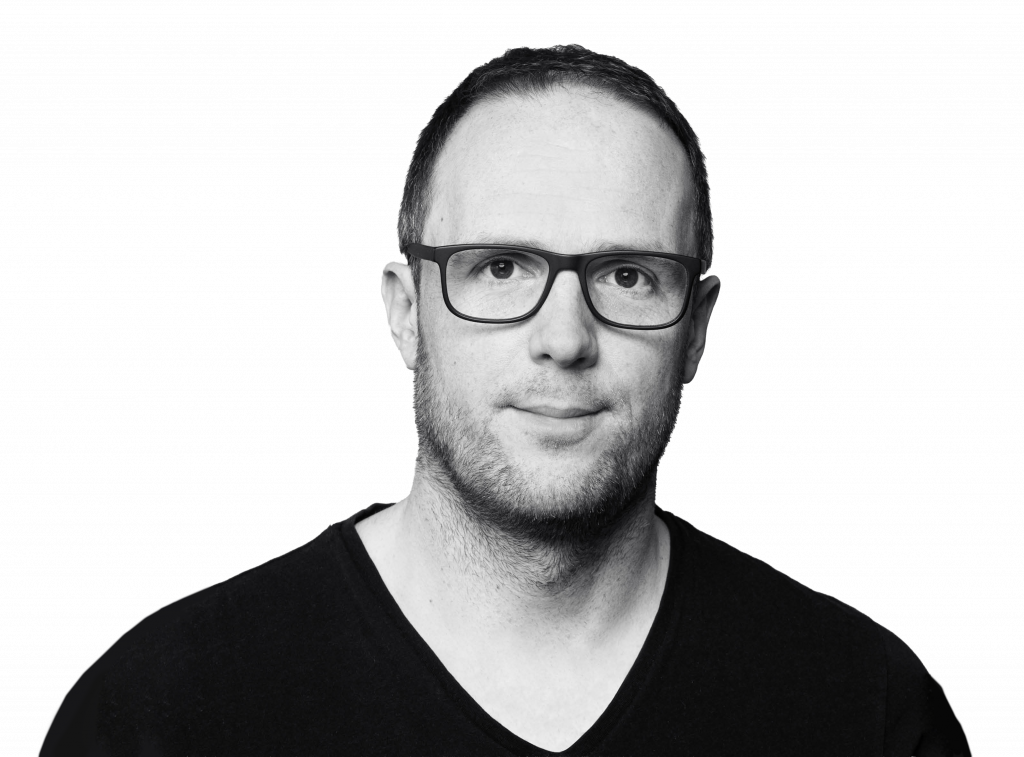 Ein Mann mit kurzen Haaren und Brille, der ein schwarzes Hemd mit V-Ausschnitt trägt, blickt nach vorne vor einem schlichten weißen Hintergrund.