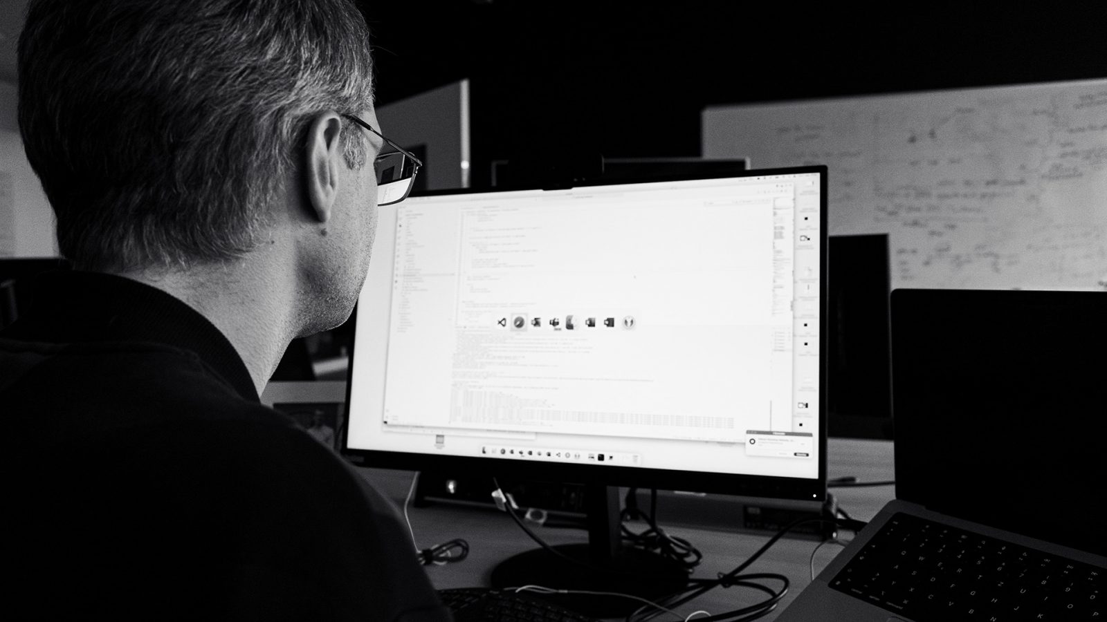 Eine Person, die eine Brille trägt, sitzt an einem Schreibtisch und arbeitet an einem Computer, auf dessen Monitor in einem schwach beleuchteten Büro Codes und Symbole angezeigt werden.