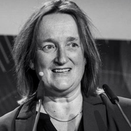 Eine Frau mit schulterlangem Haar spricht an einem Podium mit zwei Mikrofonen. Sie trägt einen Blazer und eine Halskette. Das Bild ist in schwarz-weiß gehalten.