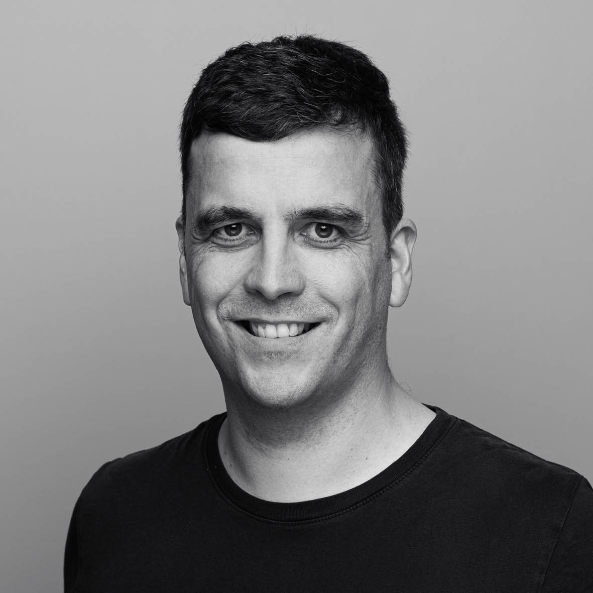 Ein Mann mit kurzen dunklen Haaren und einem schwarzen T-Shirt lächelt in die Kamera vor einem schlichten, hellen Hintergrund.