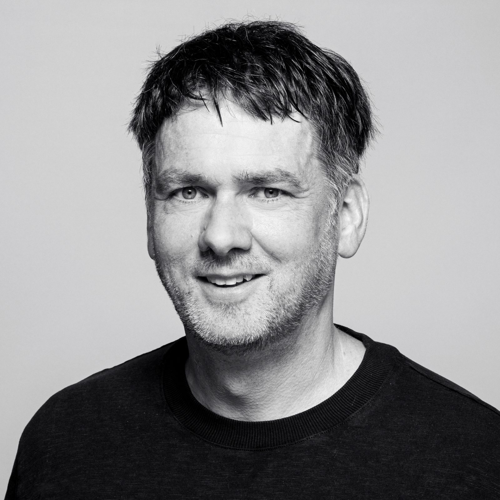 Mann mit kurzen dunklen Haaren und einem Bart, der ein dunkles T-Shirt mit Rundhalsausschnitt trägt und in die Kamera lächelt, vor einem schlichten hellen Hintergrund.