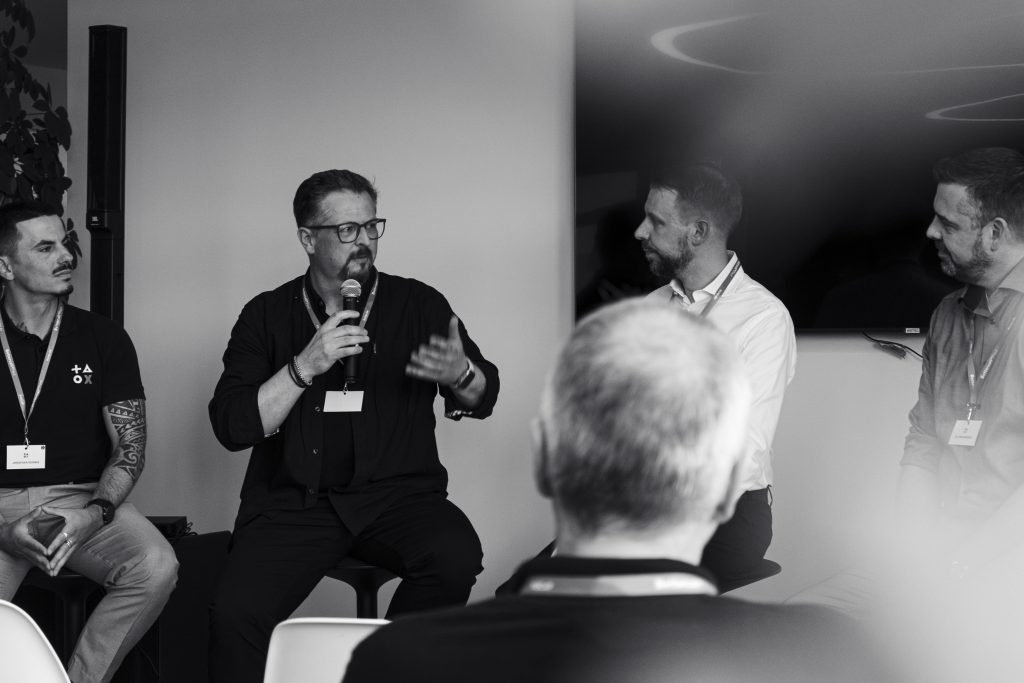 Vier Männer sitzen und sprechen vor einem Publikum; ein Mann spricht in ein Mikrofon, während die anderen zuhören. Der Rahmen scheint eine Podiumsdiskussion oder ein Seminar zu sein.