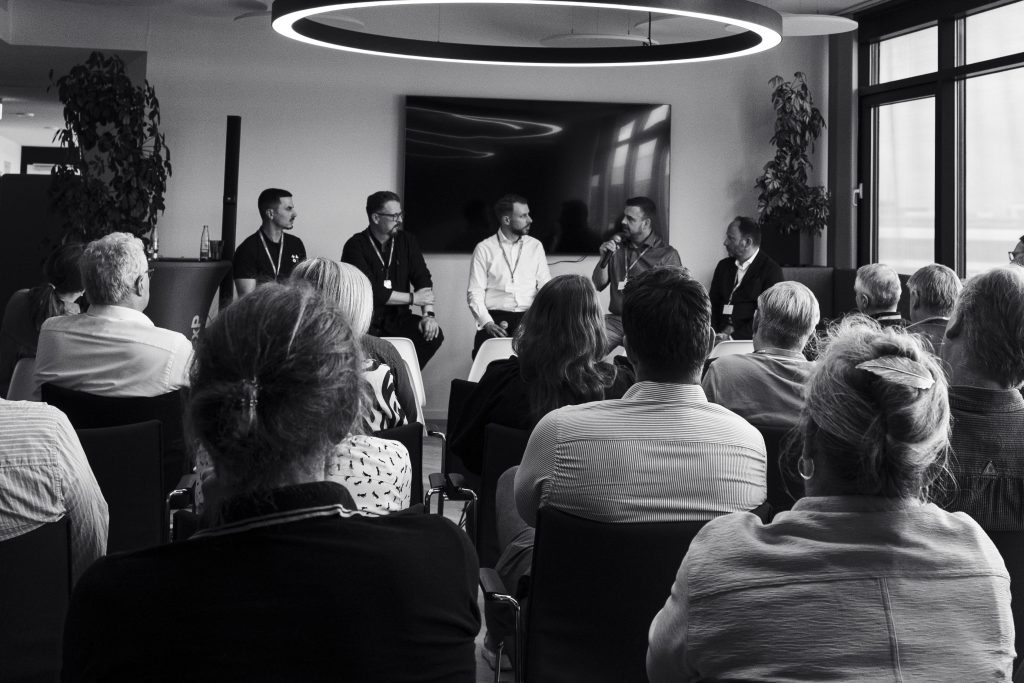 Ein fünfköpfiges Gremium spricht zu einem Publikum in einem modernen Konferenzraum; die Szene ist in Schwarz-Weiß gehalten.