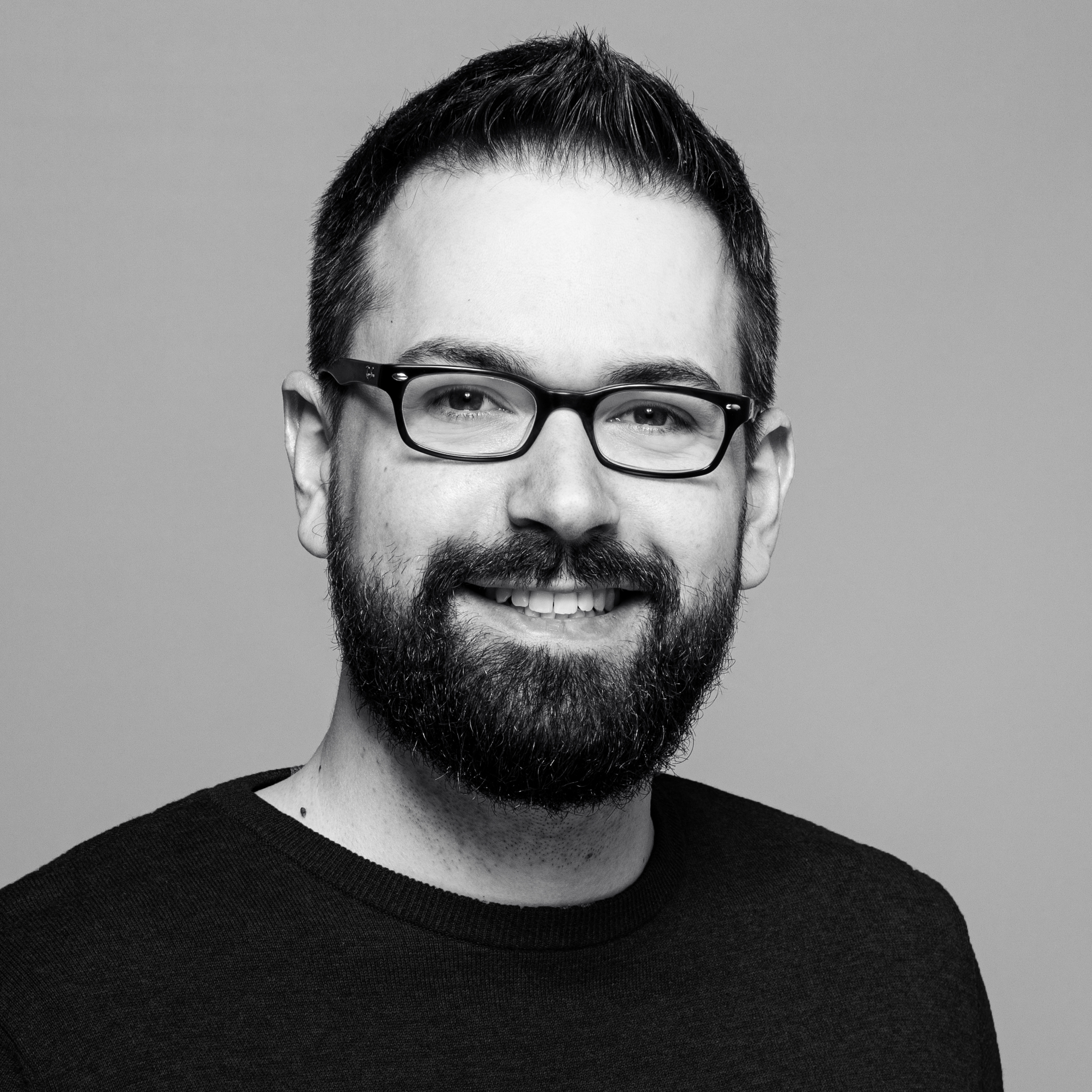 Ein Mann mit kurzen dunklen Haaren, Brille und Bart lächelt vor einem schlichten Hintergrund in einen dunklen Pullover gekleidet in die Kamera.
