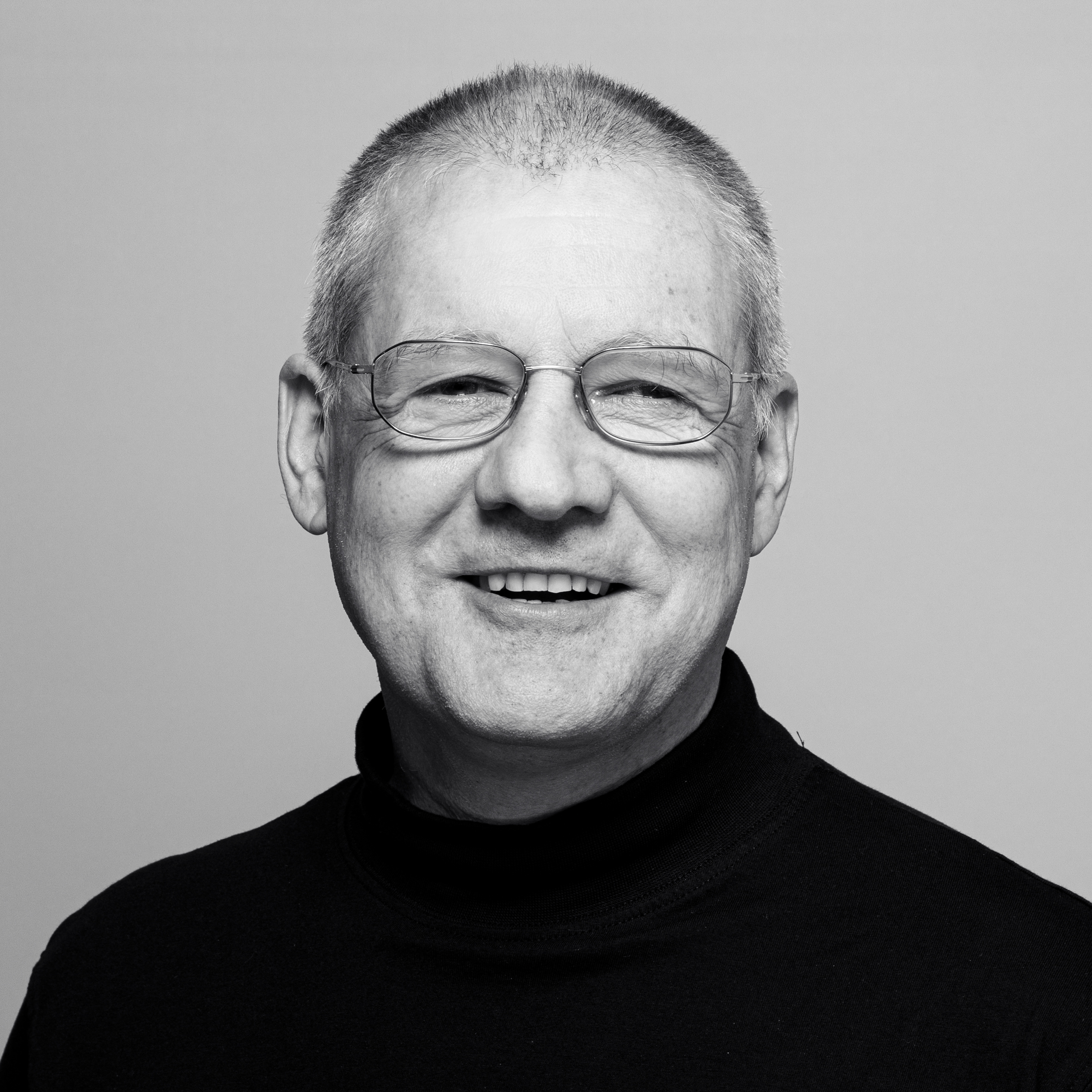 Ein Mann mittleren Alters mit kurzen grauen Haaren, Brille und einem schwarzen Rollkragenpullover lächelt vor einem schlichten Hintergrund in die Kamera.