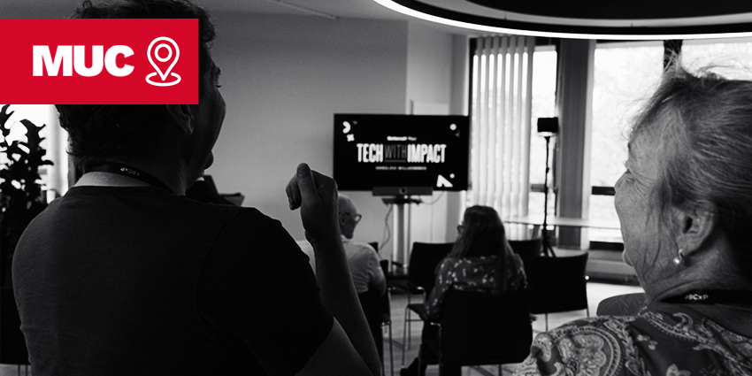 In einem Konferenzraum sitzen mehrere Personen vor einer Leinwand, auf der „TECH IMPACT“ zu lesen ist. Im Vordergrund unterhalten sich zwei Personen. In der oberen linken Ecke ist das rote „MUC“-Logo eingeblendet.