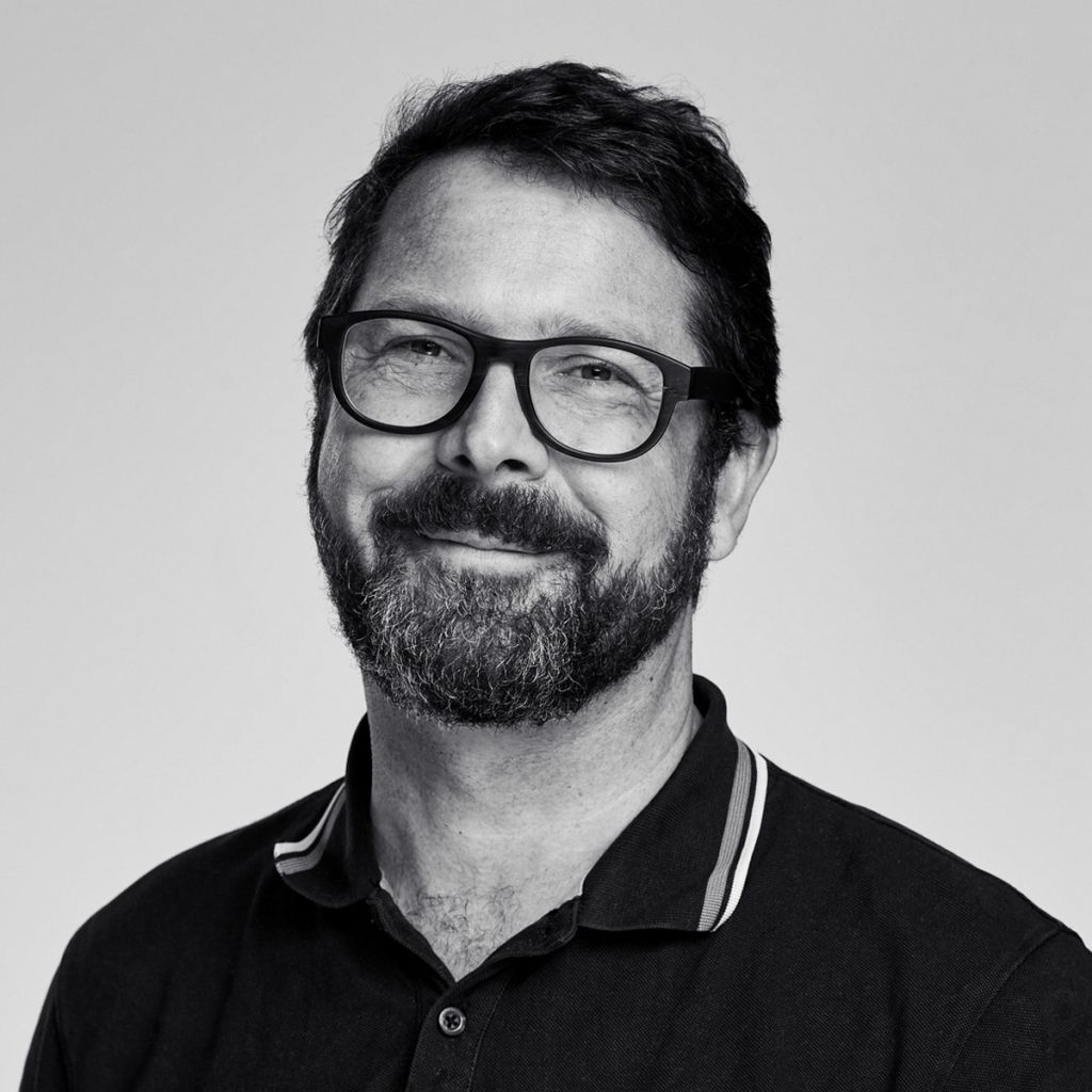 Ein Mann mit Brille und Bart, der ein dunkles Hemd mit Kragen trägt und vor einem schlichten, hellen Hintergrund leicht in die Kamera lächelt.