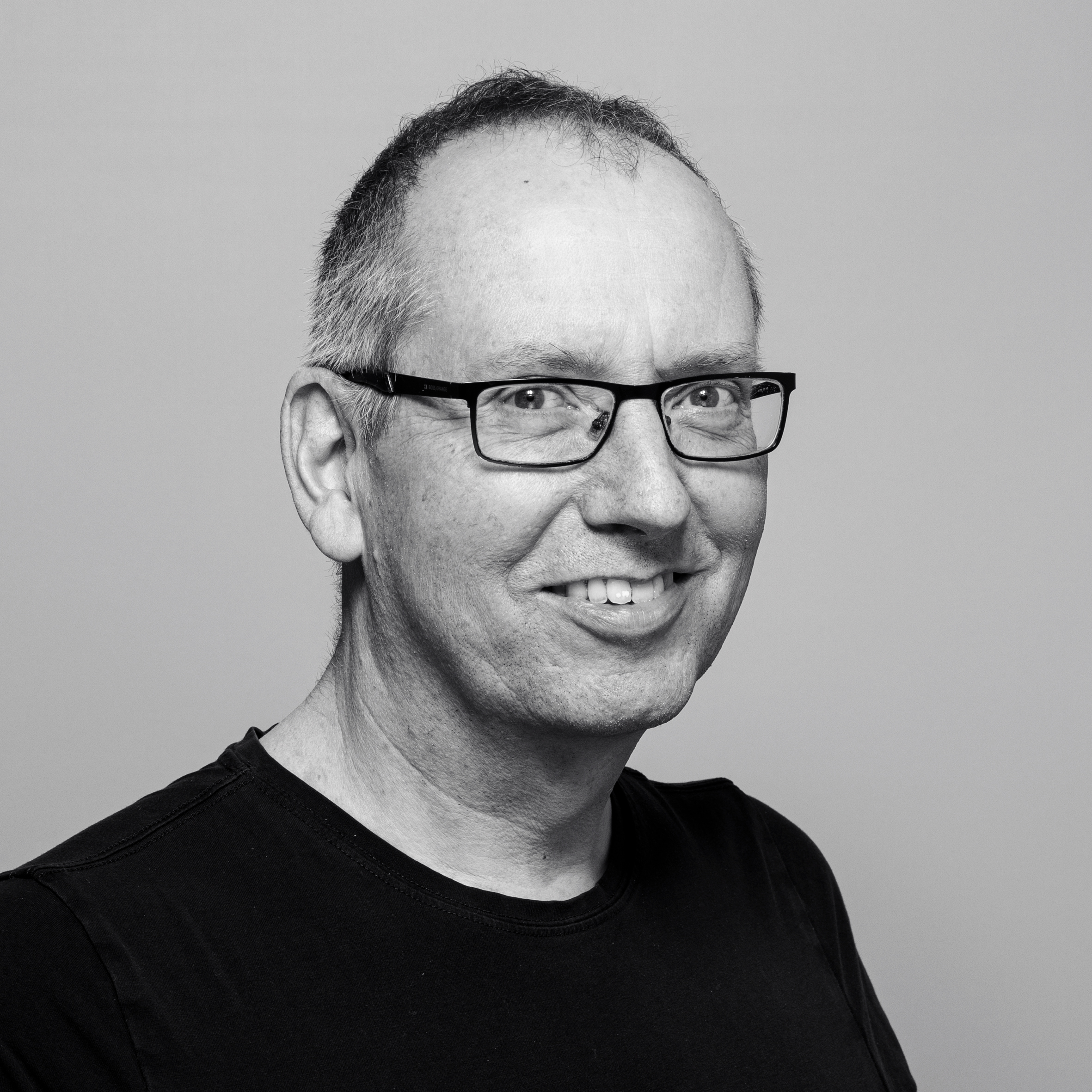 Ein Mann mittleren Alters mit Brille, kurzen Haaren und einem schwarzen Hemd lächelt vor einem schlichten, hellen Hintergrund in die Kamera.